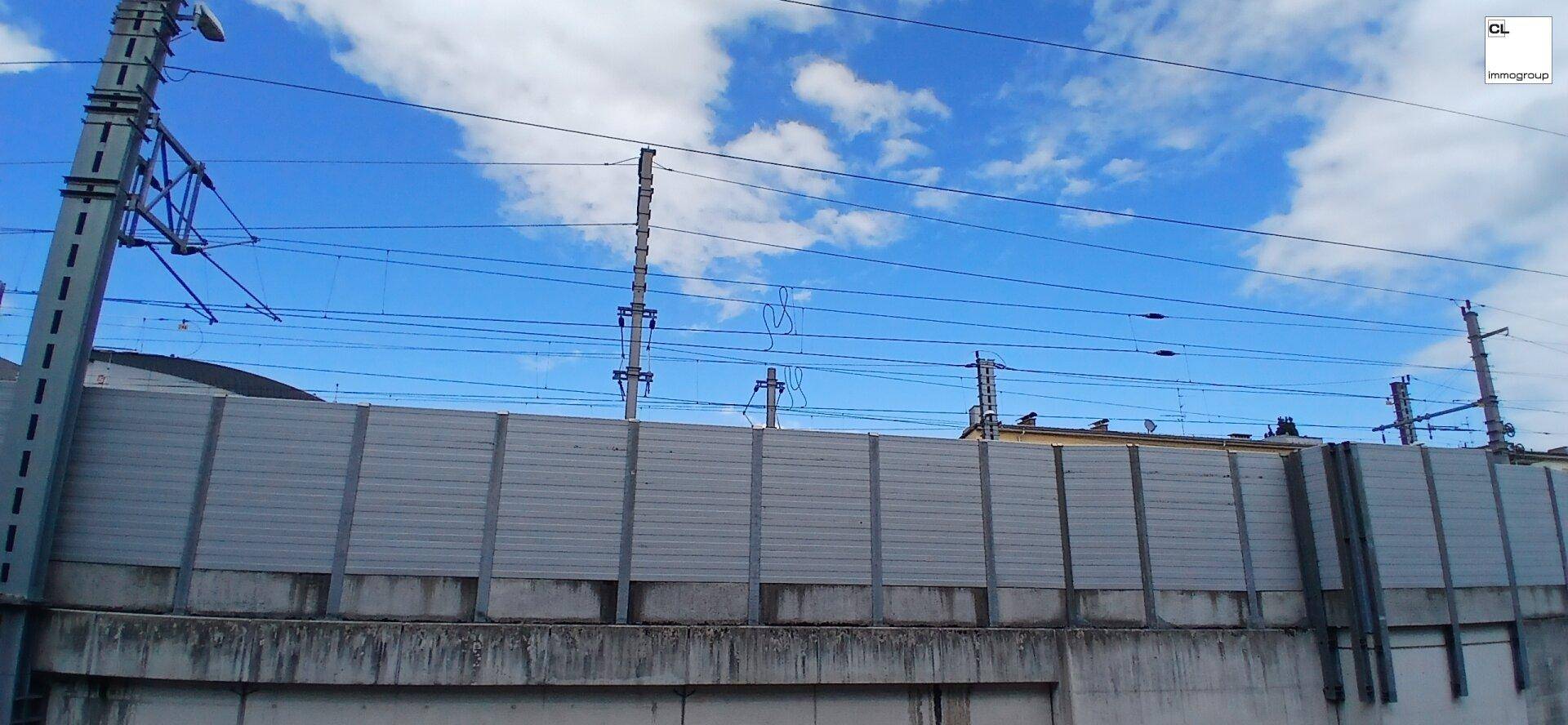 Weitblick auf Bahngleise mit Oberleitungen und einer hohen Lärmschutzwand unter blauem Himmel.