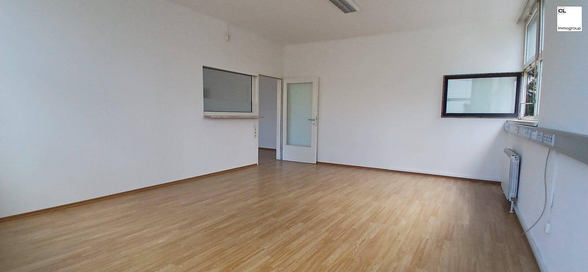 Leerer Büroraum mit Holzboden, weißen Wänden und zwei Fensteröffnungen, die Licht hereinlassen.