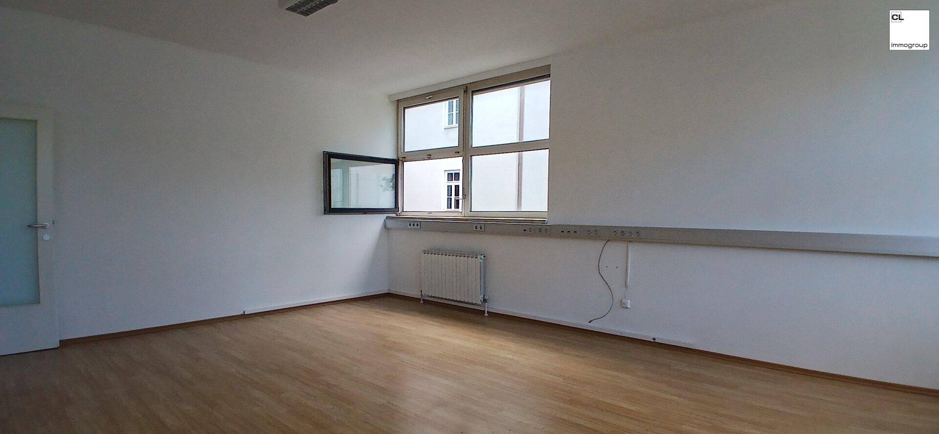 Heller Büroraum mit Holzboden, weiß gestrichenen Wänden und einem großen Fenster mit Heizkörper.