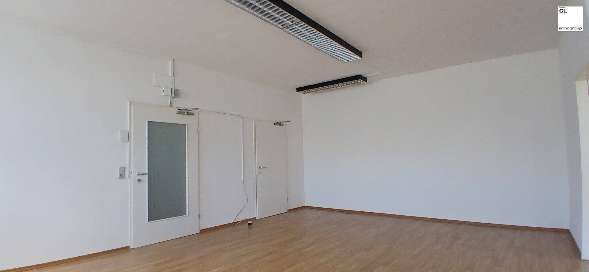 Geräumiger Büroraum mit Holzboden, weißen Wänden und zwei Türen, ausgestattet mit Deckenbeleuchtung.