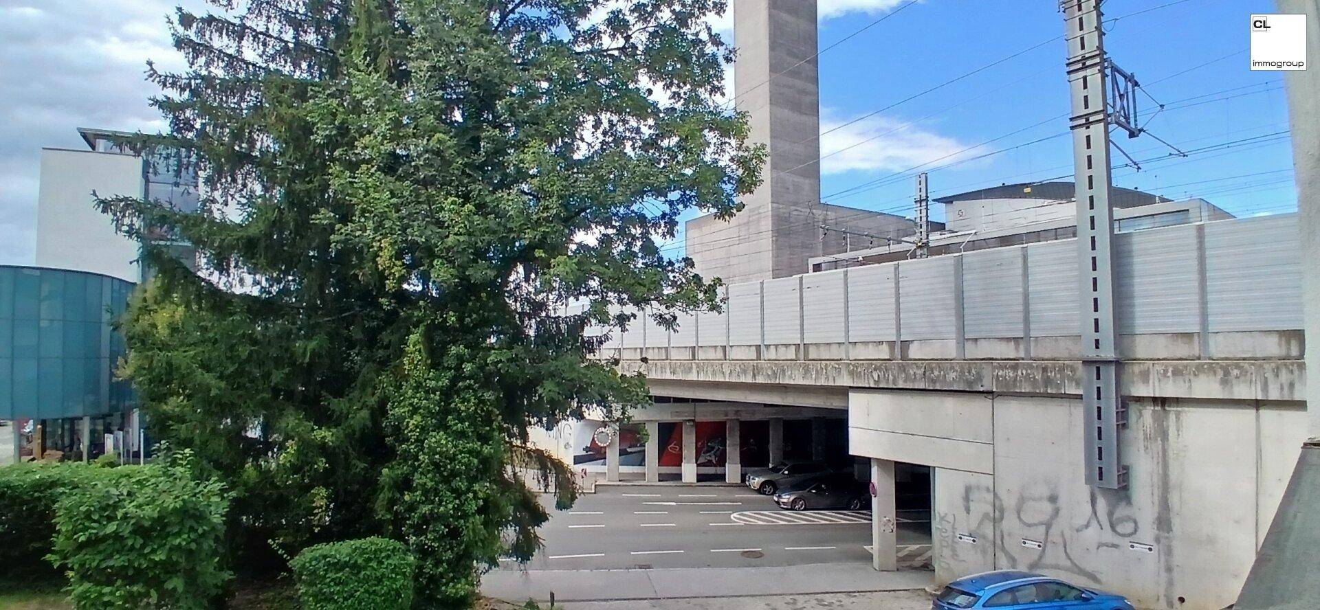 Außenansicht mit großem Baum, Gebäude, Eisenbahnbrücke und einem Parkplatz darunter.