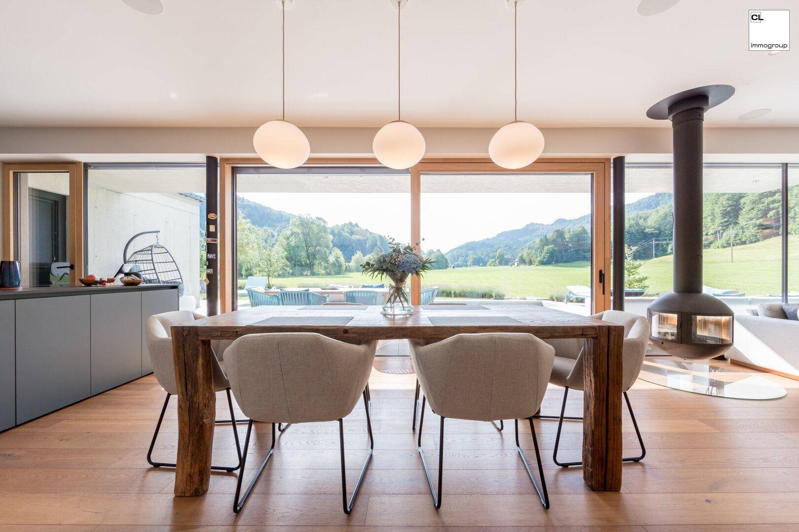 Heller Wohn-Essbereich mit Holztisch, modernem Kamin und Panoramafenstern mit Bergblick.