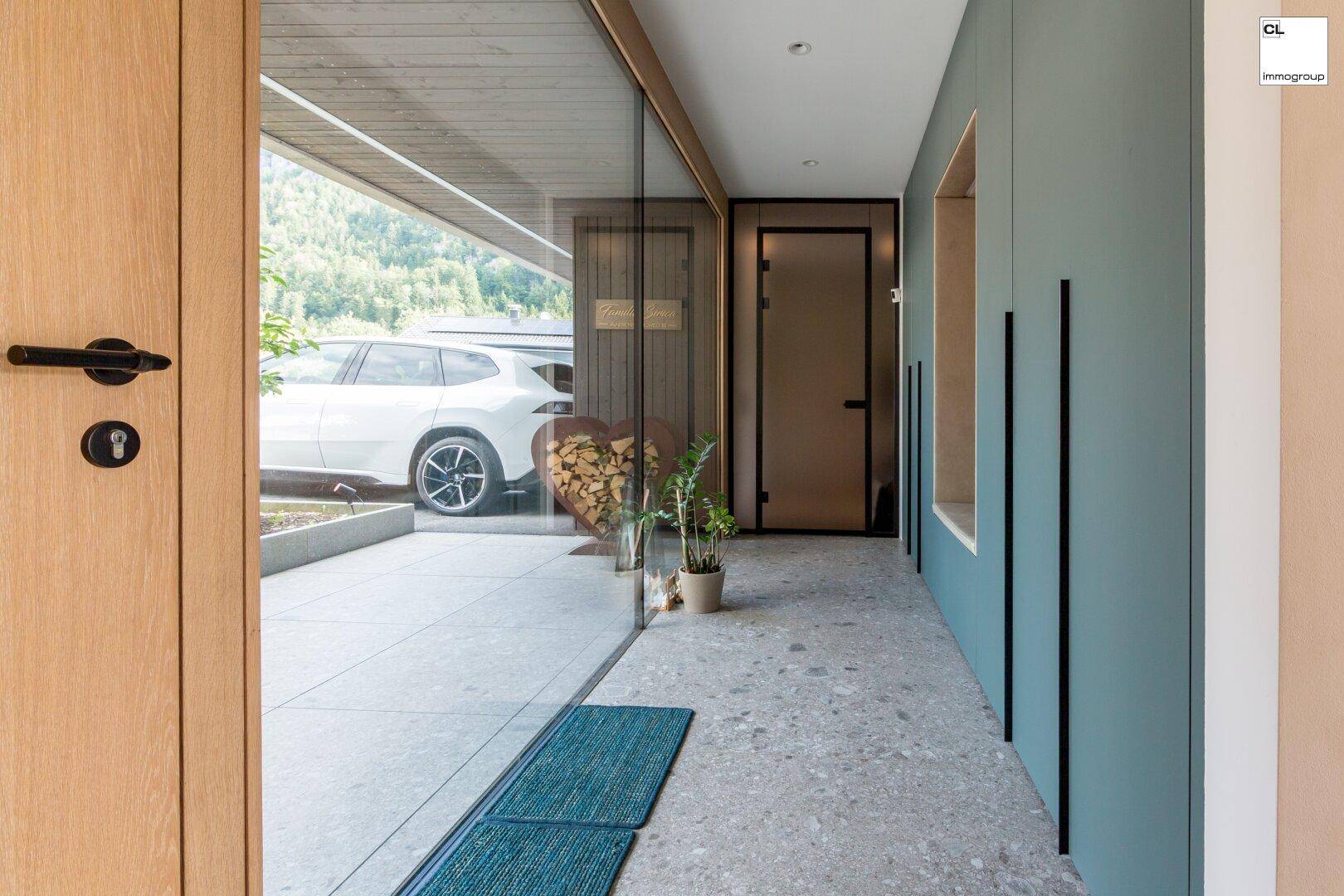 Heller Eingangsbereich mit Holztür, Glasfront und Blick auf den Carport mit Fahrzeug.