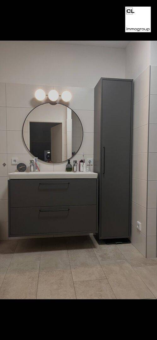 Modernes Badezimmer mit grauem Waschtischunterschrank, rundem Spiegel und hellen Fliesen.