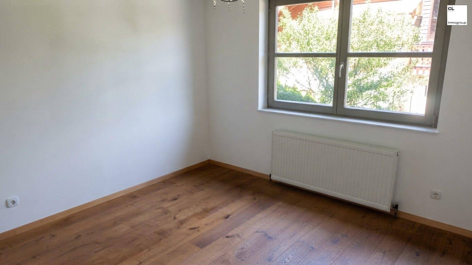 Helles Zimmer mit Holzboden, weiß gestrichenen Wänden und einem großen Fenster mit Blick ins Grüne.