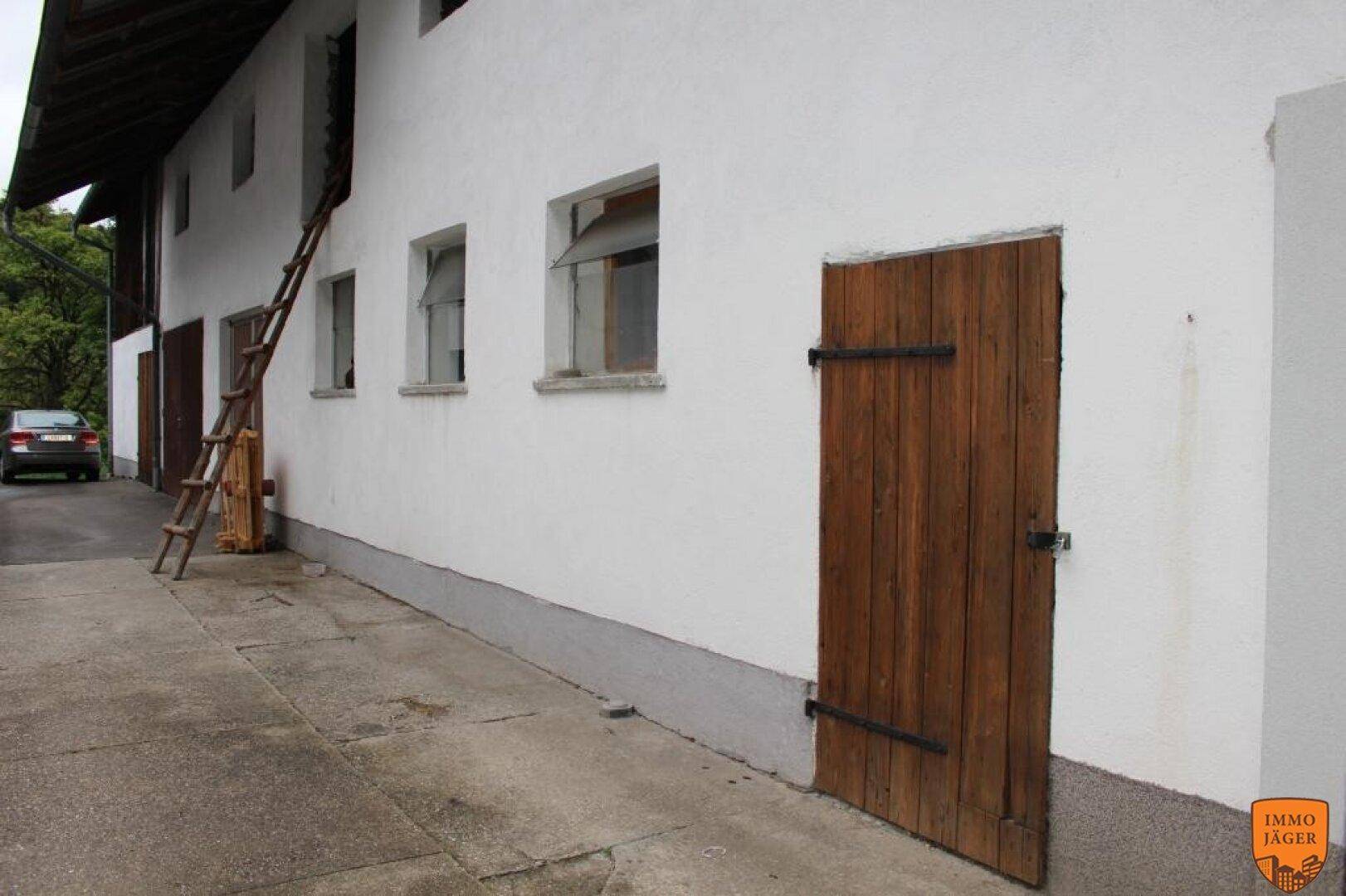 Seitenansicht eines älteren Gebäudes mit weißer Fassade, Holztür und mehreren Fenstern, daneben eine Auffahrt.