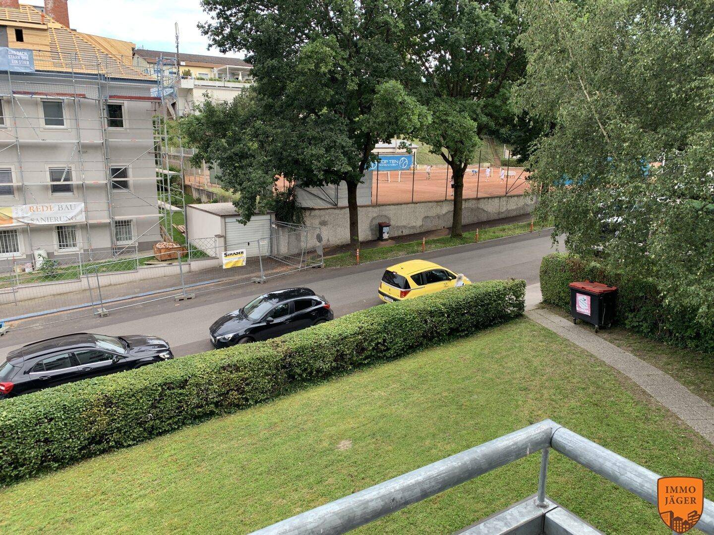 Balkon mit Blick auf eine Straße, Grünflächen und einen Tennisplatz in der Ferne.