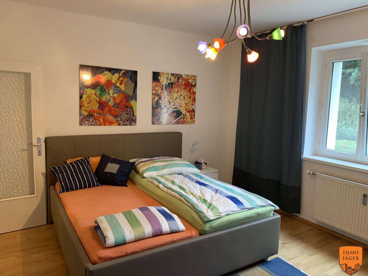 Schlafzimmer mit Doppelbett, bunten Bildern und einem Fenster mit Blick ins Grüne.