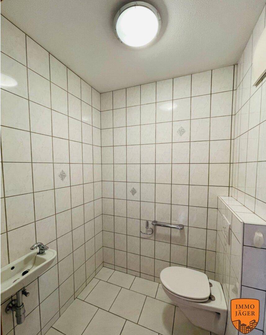 Kleines Gäste-WC mit weißen Fliesen, Toilette, kleinem Waschbecken und Haltegriff.