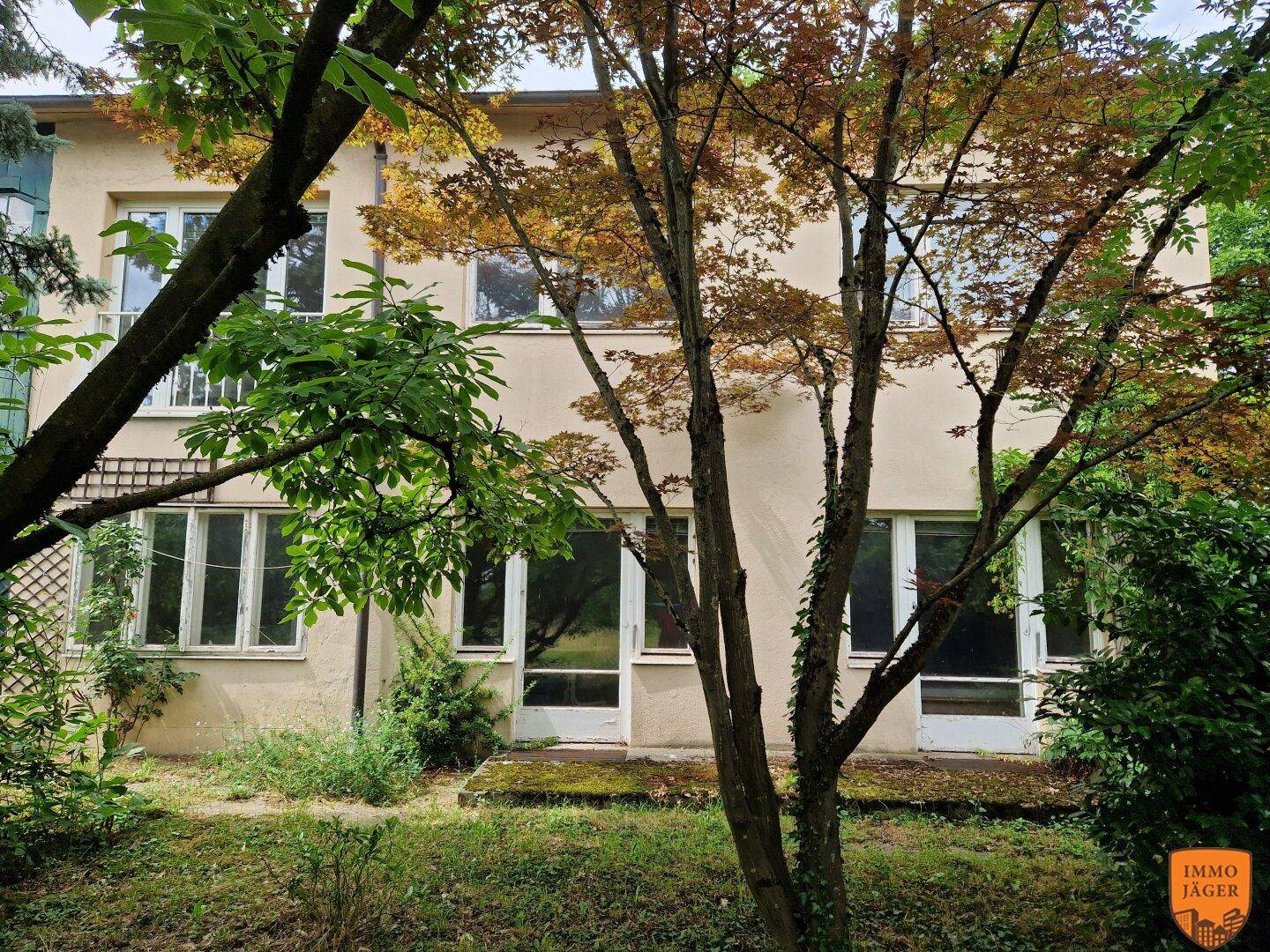 Fassade eines Hauses mit großen Fenstern im Erdgeschoss, umgeben von Bäumen und Sträuchern.