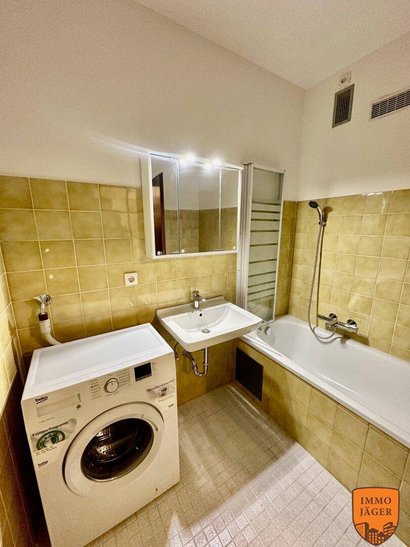 Badezimmer mit Badewanne, Waschbecken, Spiegelschrank und Waschmaschine, gefliest in Gelbtönen.