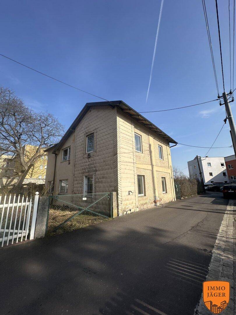 Seitenansicht des Wohnhauses mit heller Fassade und Zaun, an einer schmalen Straße gelegen.