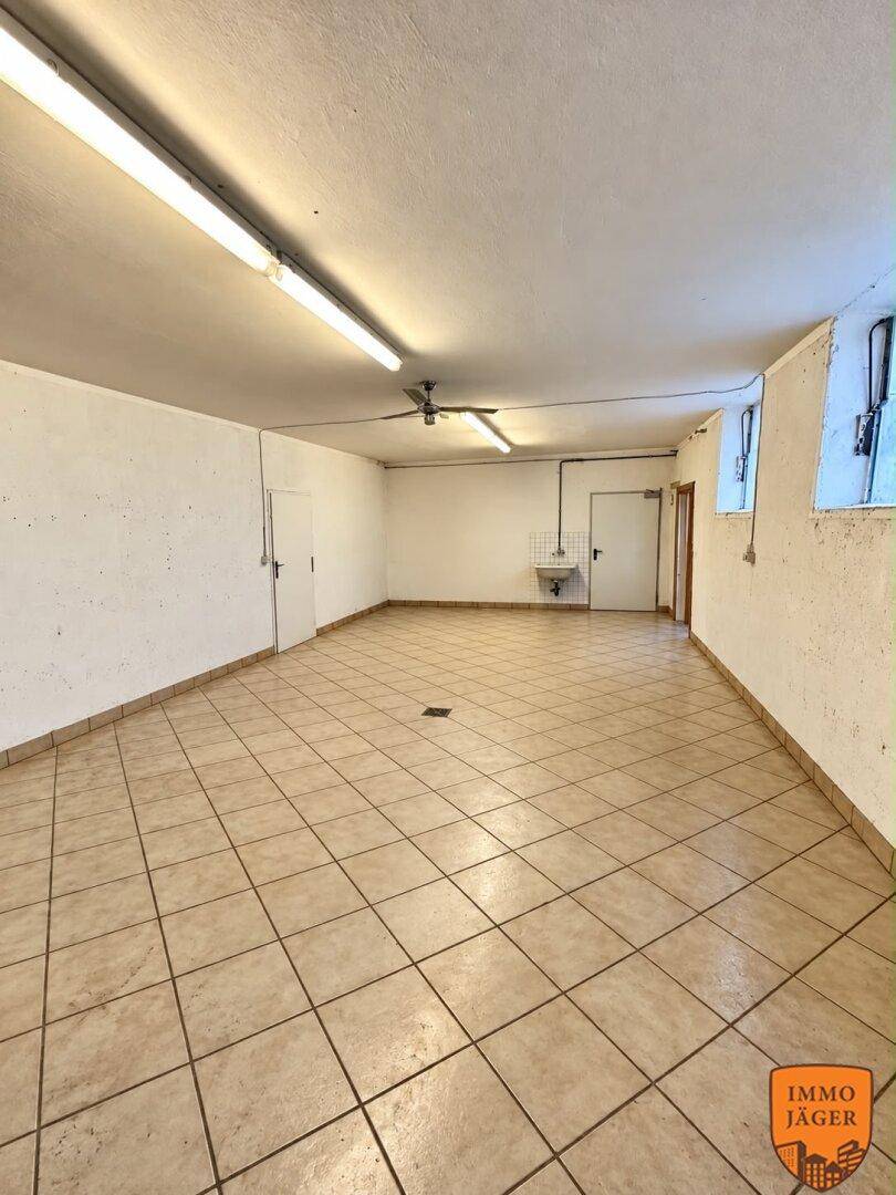 Funktionaler Kellerraum mit gefliestem Boden, Waschbecken und zwei Türen, gut beleuchtet durch Leuchtstoffröhren.