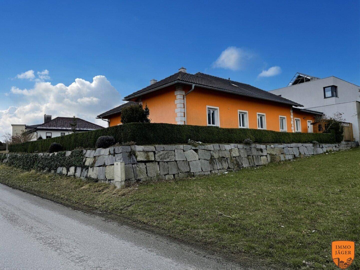 Orangefarbenes Einfamilienhaus mit Natursteinmauer und hoher Hecke am Straßenrand.