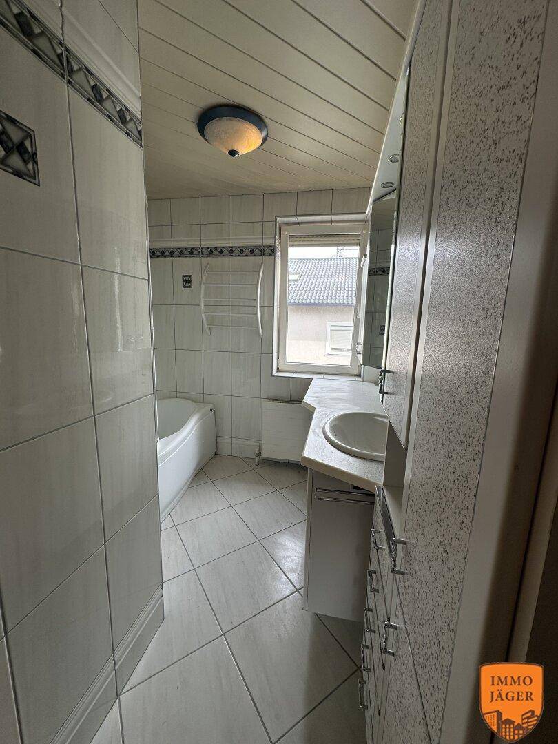 Helles Badezimmer mit weißen Fliesen, Badewanne, Einzelwaschbecken und einem Fenster für Tageslicht.