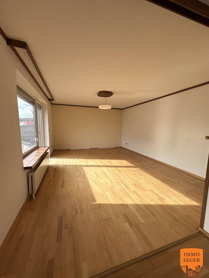 Helles, leeres Wohnzimmer mit Parkettboden und großem Fenster, das viel Tageslicht hereinlässt.