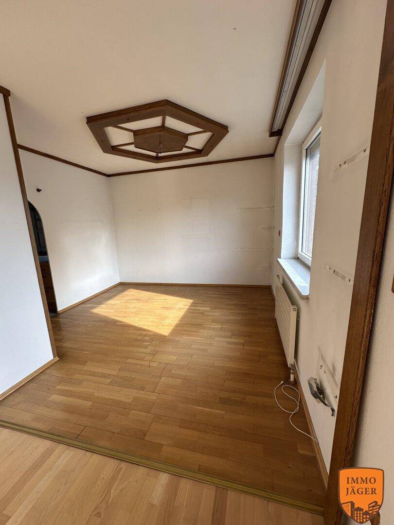 Leeres Wohnzimmer mit Parkettboden, dekorativer Holzdecke und großem Fenster, das viel Licht spendet.