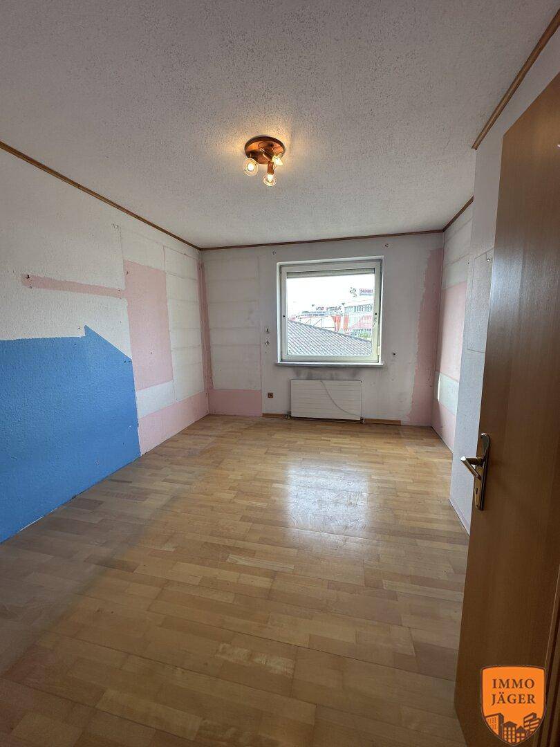 Schlafzimmer mit Parkettboden, teilweise entfernter Tapete und einem Fenster mit Blick nach draußen.