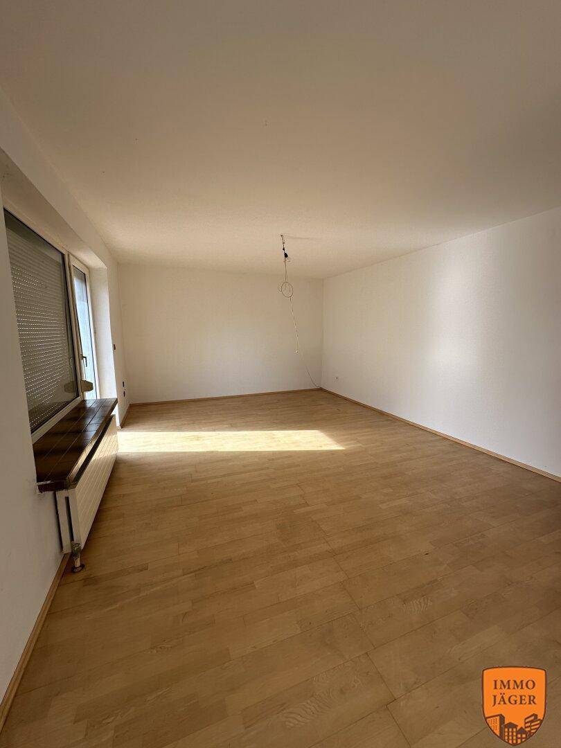 Helles, leeres Wohnzimmer mit Parkettboden und großem Fenster mit Jalousien, das viel Tageslicht hereinlässt.
