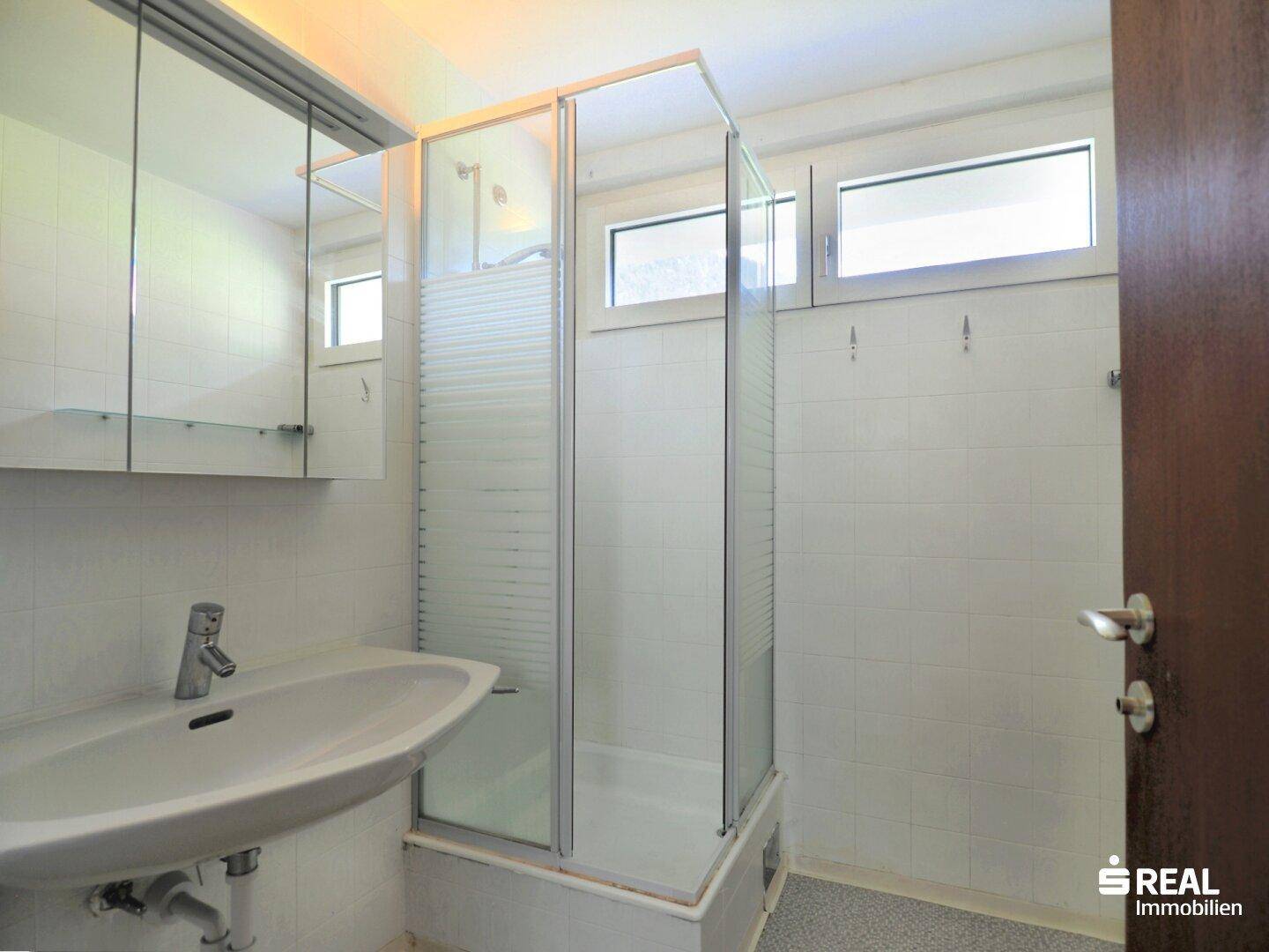 Badezimmer mit Duschkabine, Einzelwaschbecken und Fenster, gefliest in hellem Weiß.