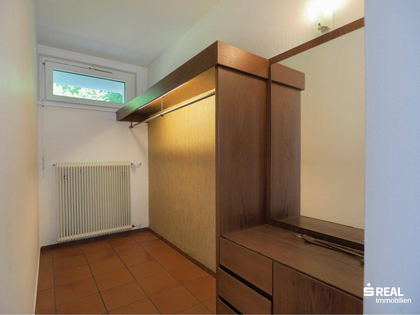 Begehbarer Kleiderschrank mit Holzregalen, Fenster und Terrakotta-Fliesenboden.