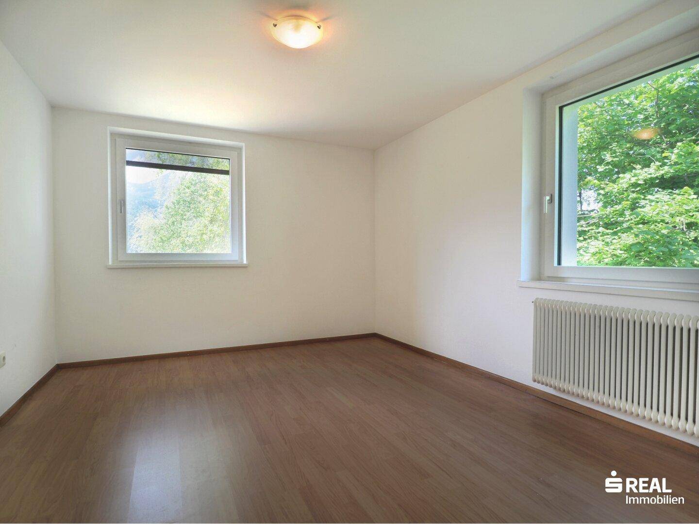 Schlafzimmer mit hellem Holzboden und zwei Fenstern, die viel Tageslicht hereinlassen.