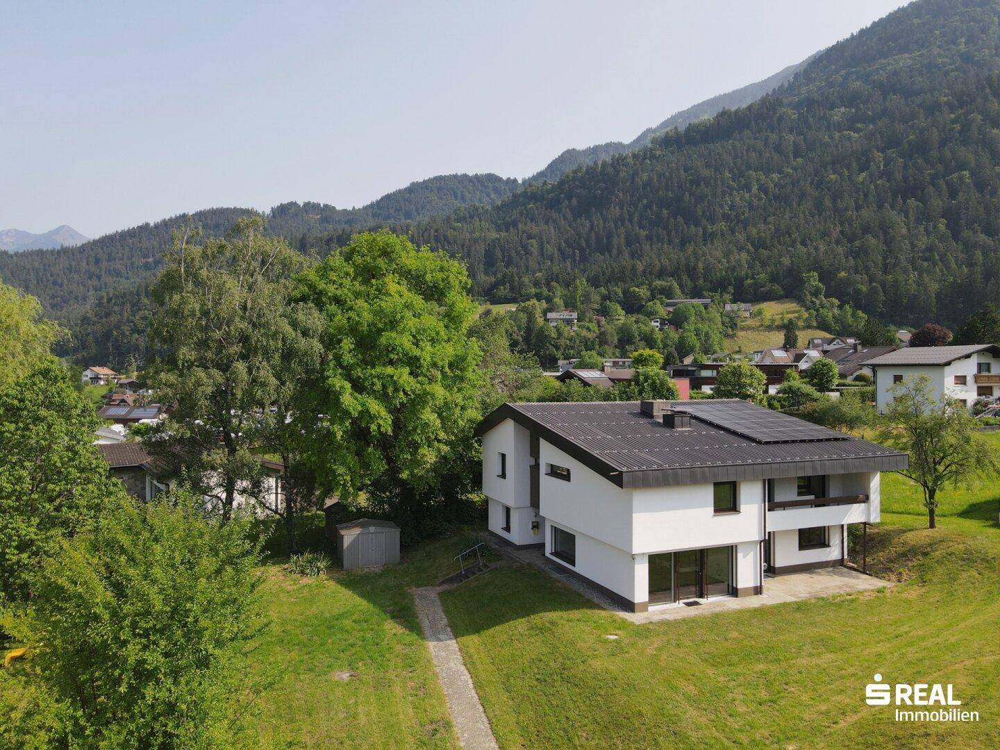 Freistehendes Haus mit Photovoltaik auf dem Dach, umgeben von viel Grün und Bergen im Hintergrund.