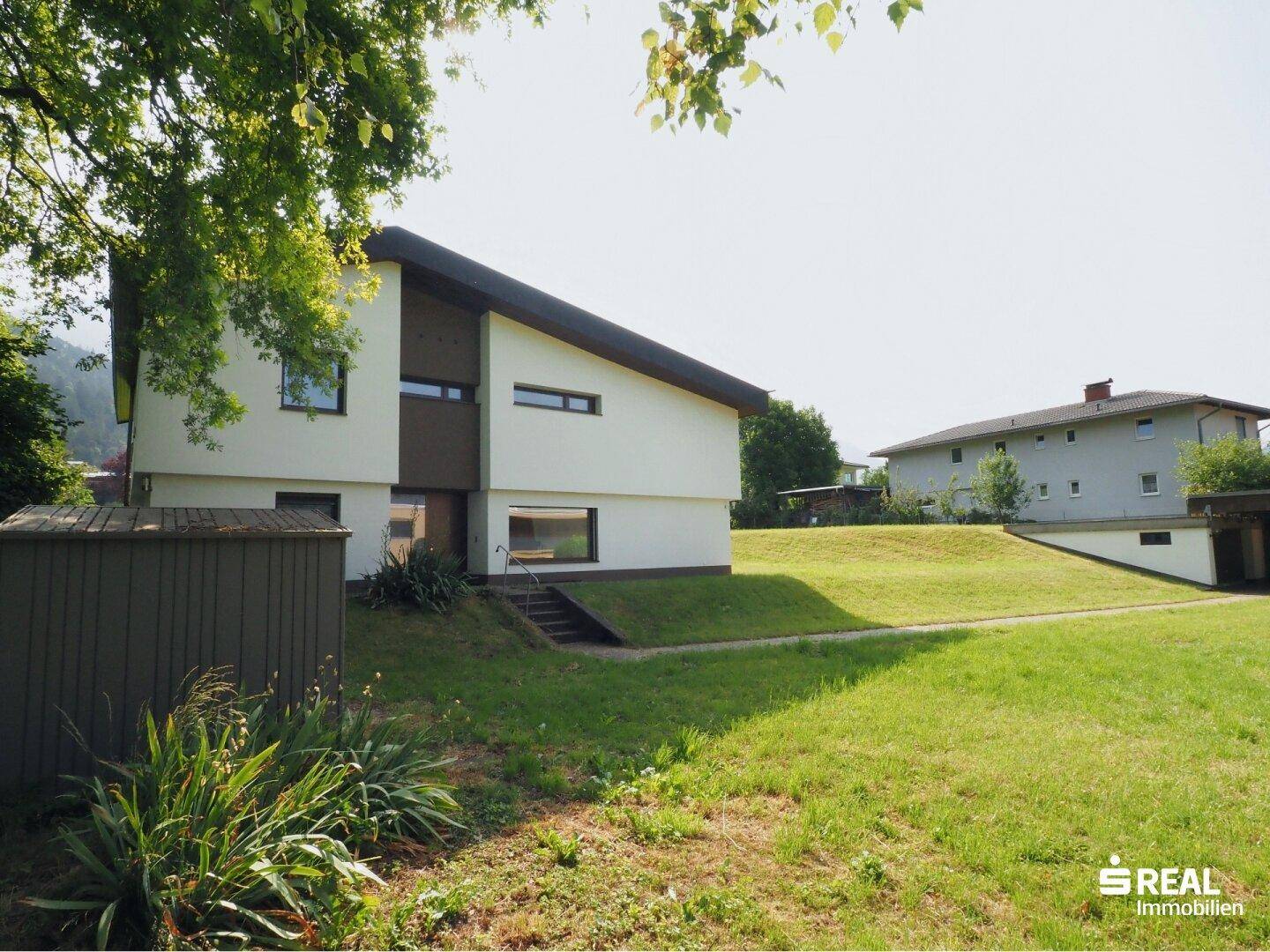 Seitenansicht des Hauses mit Gartenhütte und weitläufiger Rasenfläche in sonniger Lage.