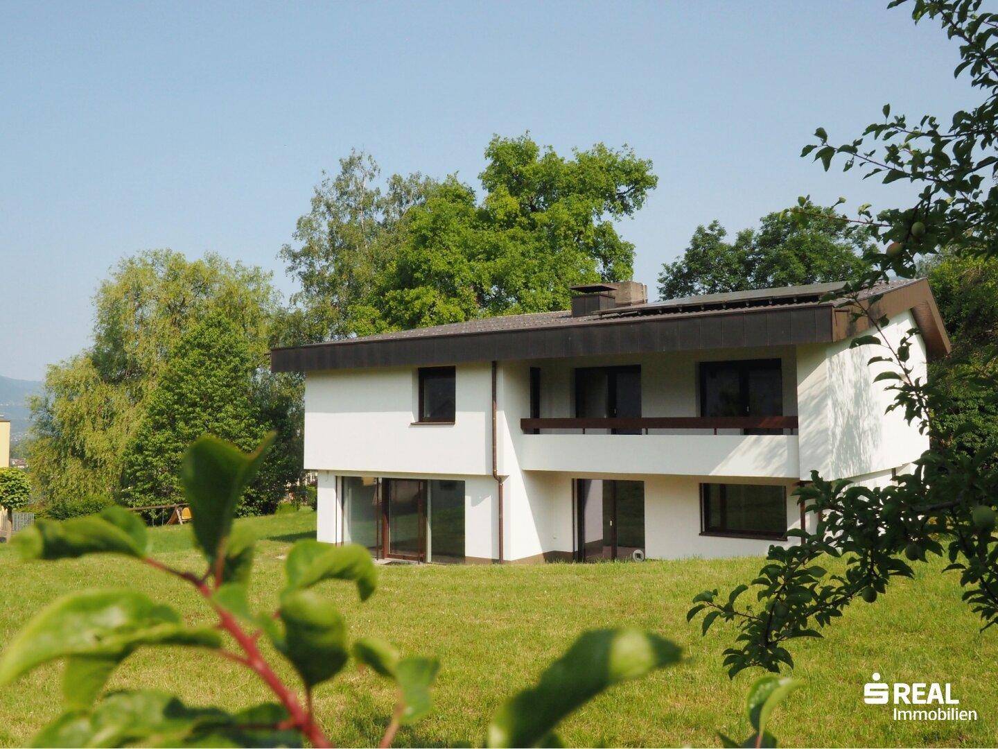 Freistehendes Haus mit weißer Fassade und dunklem Dach, umgeben von üppigem Grün.