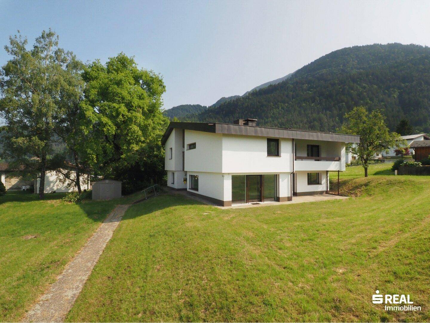 Modernes Einfamilienhaus mit Photovoltaikanlage auf dem Dach, umgeben von viel Grün und Bergen.