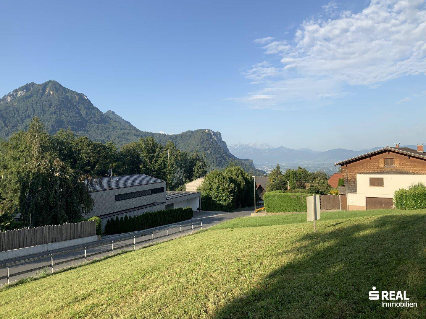 Weitläufige grüne Landschaft mit Häusern, einer Straße und beeindruckendem Bergpanorama unter blauem Himmel.