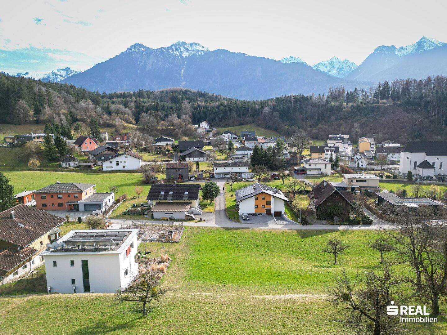 Weitläufige Luftaufnahme eines Dorfes mit grünen Feldern und majestätischen Bergen im Hintergrund.