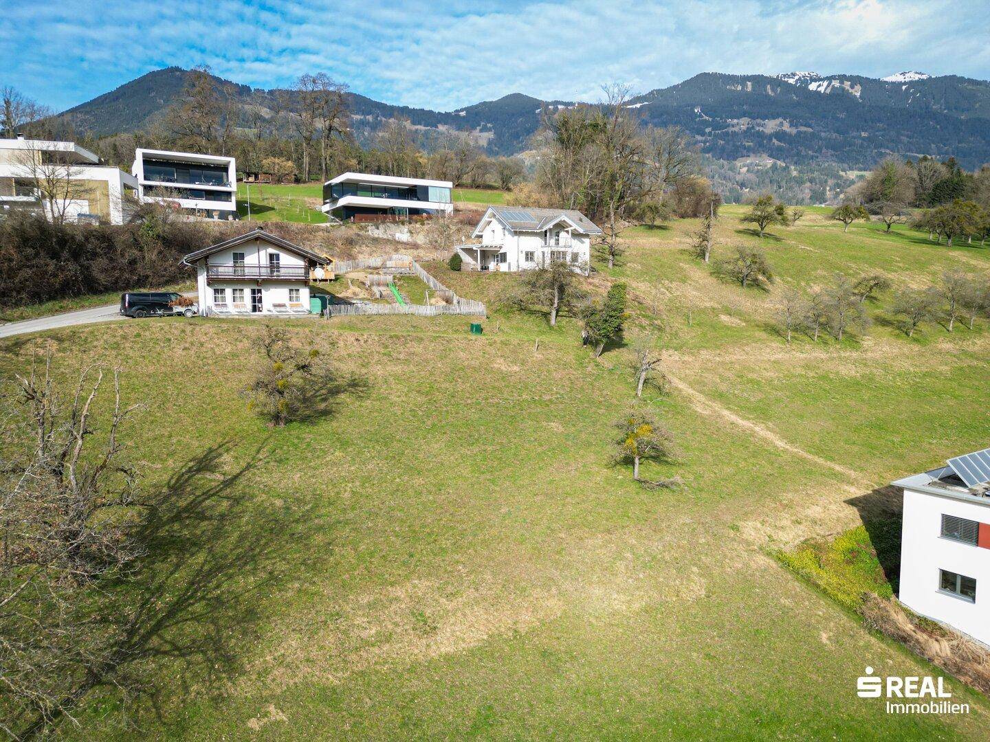 Luftaufnahme einer Wohnsiedlung am Hang, eingebettet in eine grüne Landschaft mit Bergblick.