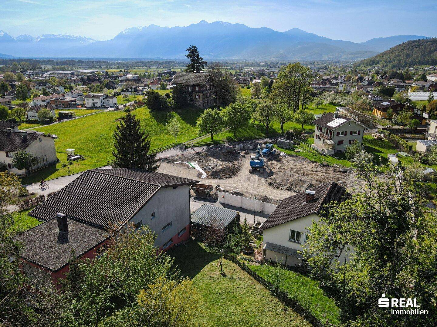 Luftaufnahme einer ländlichen Siedlung mit Baustellenbereich und Bergpanorama im Hintergrund.