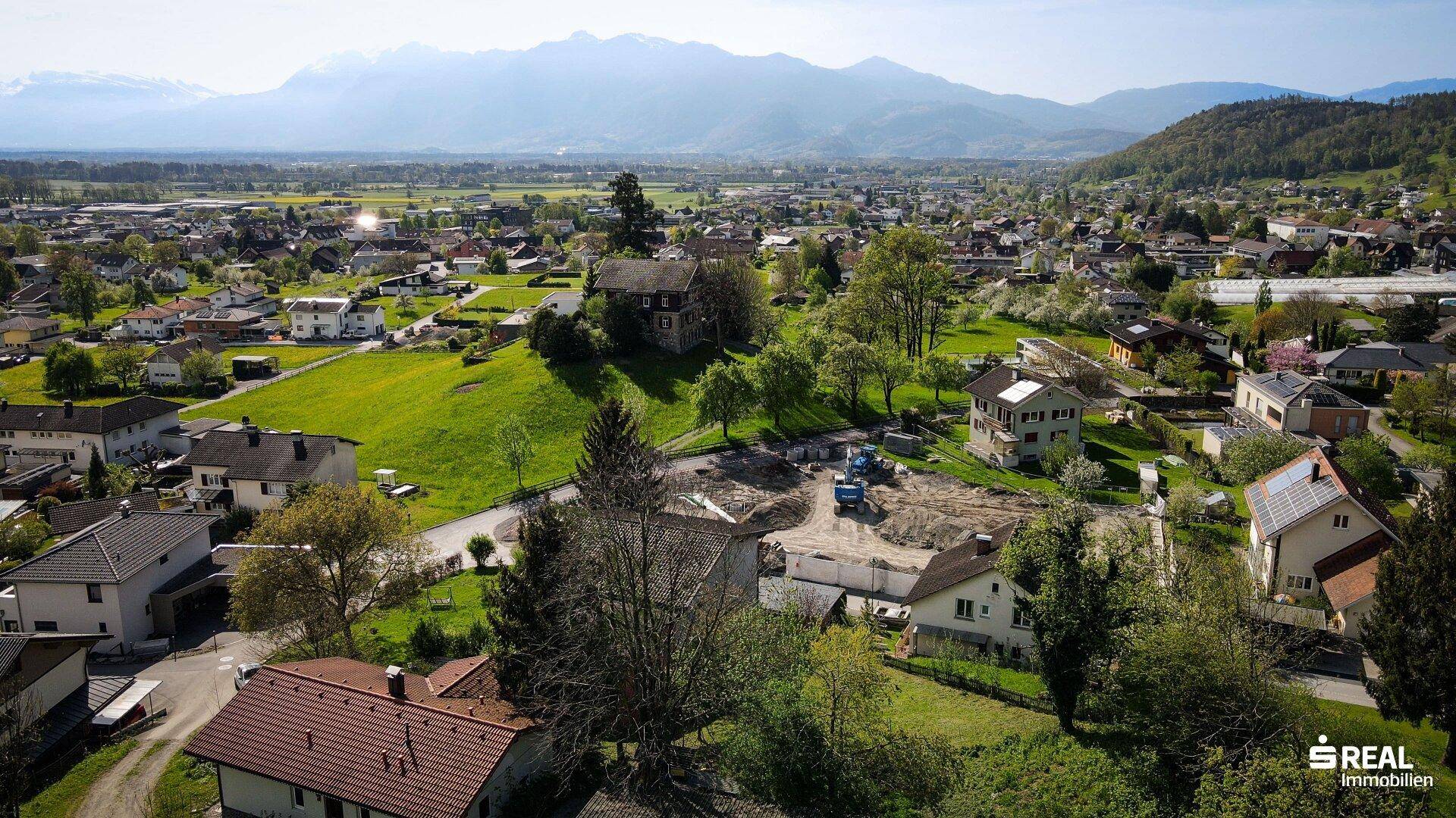Weitläufige Luftaufnahme einer Siedlung mit grünen Hügeln und majestätischen Bergen im Hintergrund.