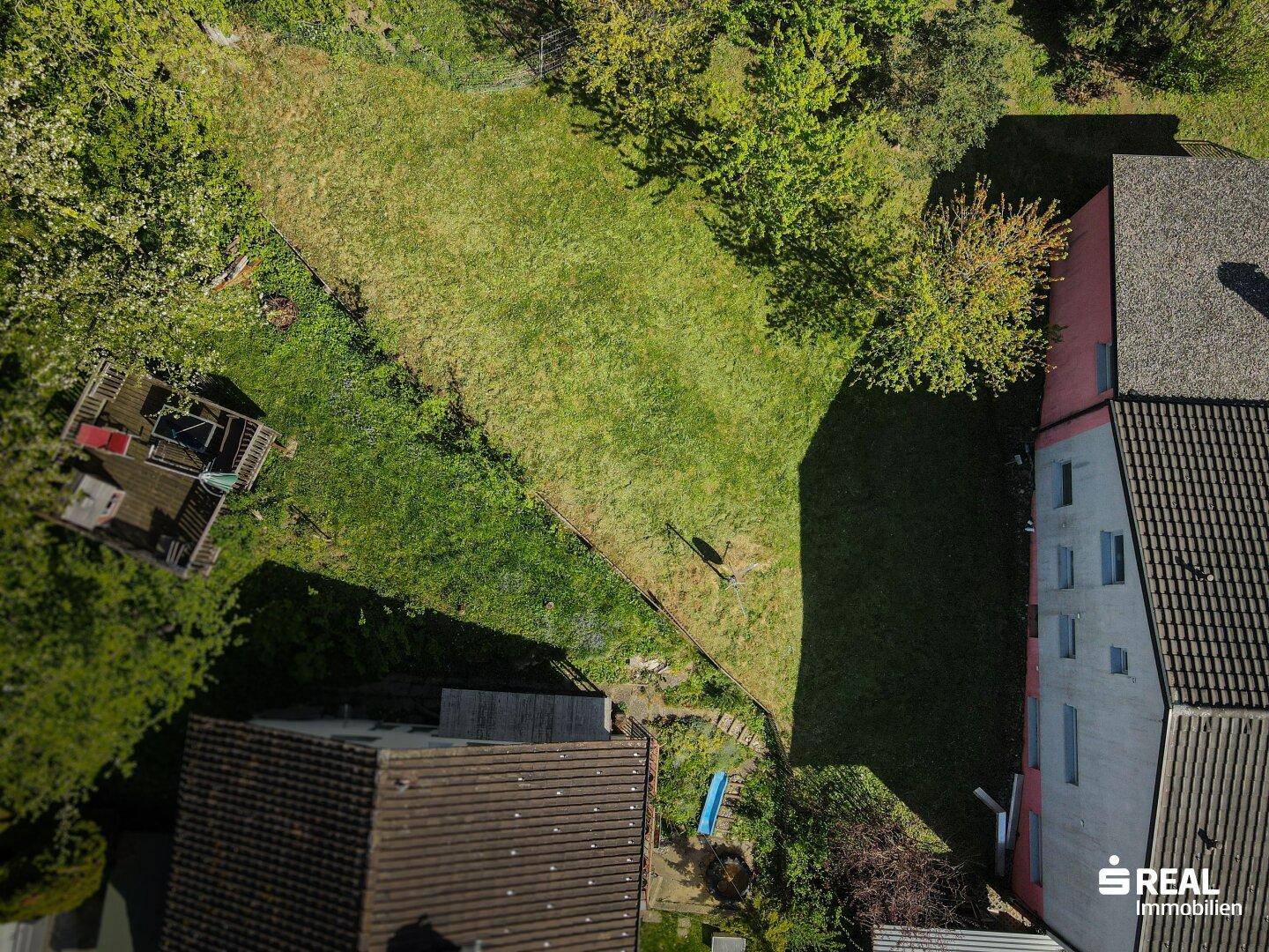 Luftaufnahme eines Gartens mit Rasenfläche, Holzterrasse und angrenzendem Wohnhaus.