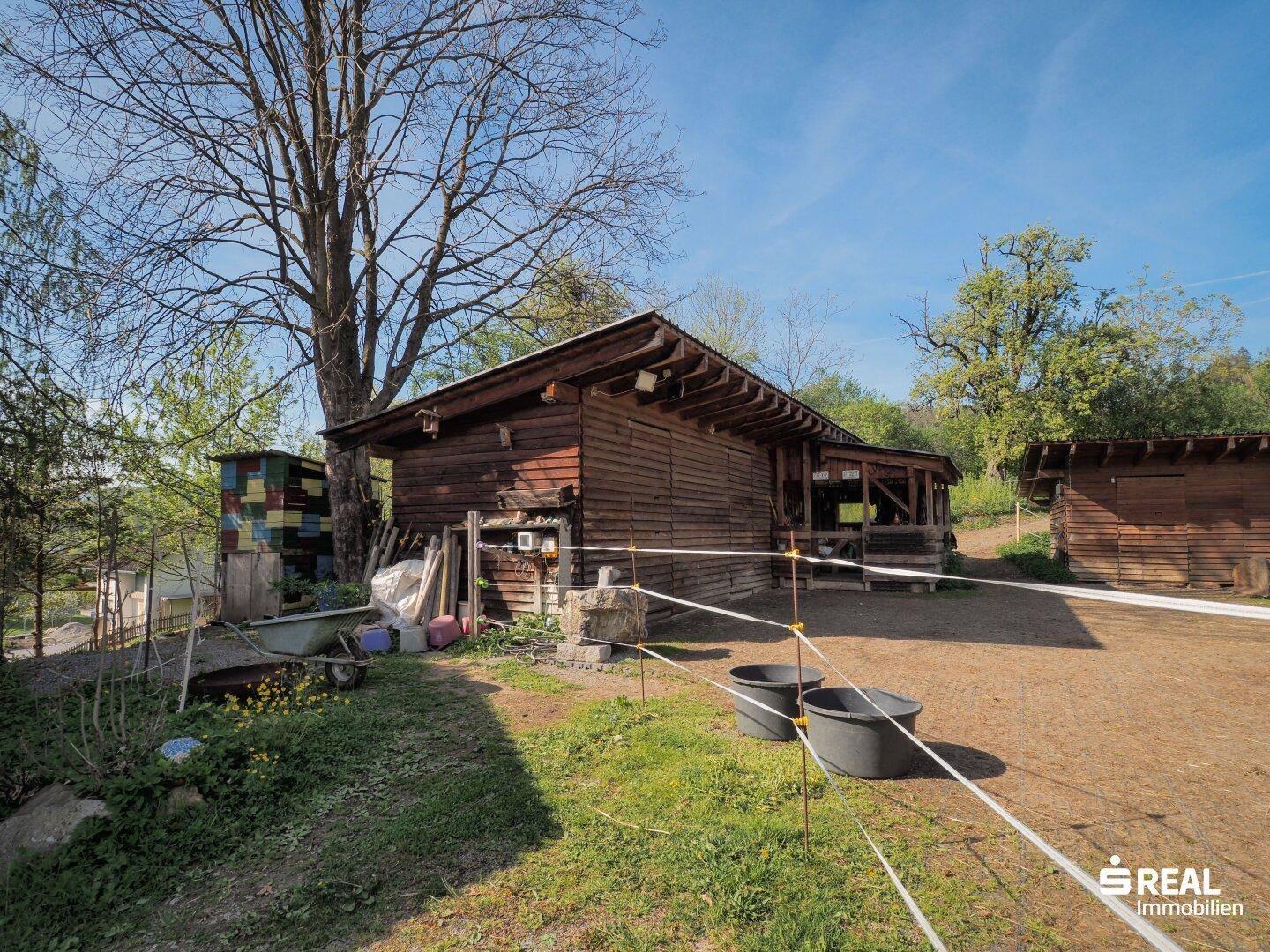 Rustikales Holzgebäude mit Vordach und umzäuntem Bereich, umgeben von Grünflächen.