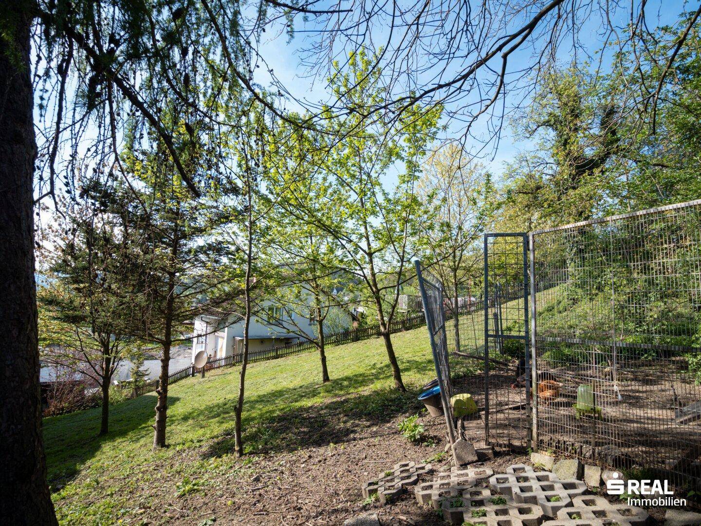 Eingezäunter Gartenbereich mit Pflastersteinen und Blick auf ein Wohnhaus in der Ferne.