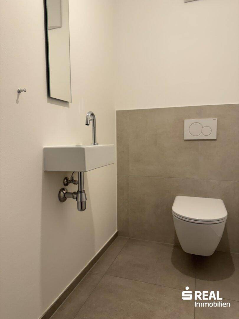 Modernes Gäste-WC mit Fliesenboden, wandhängendem WC und kleinem Waschbecken mit Spiegel.