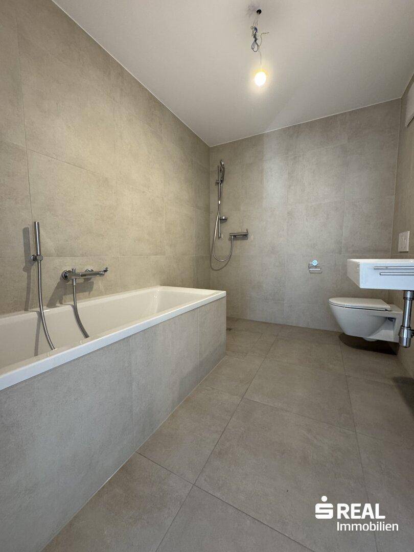 Modernes Badezimmer mit grauen Fliesen, Badewanne, Dusche und wandhängendem WC.