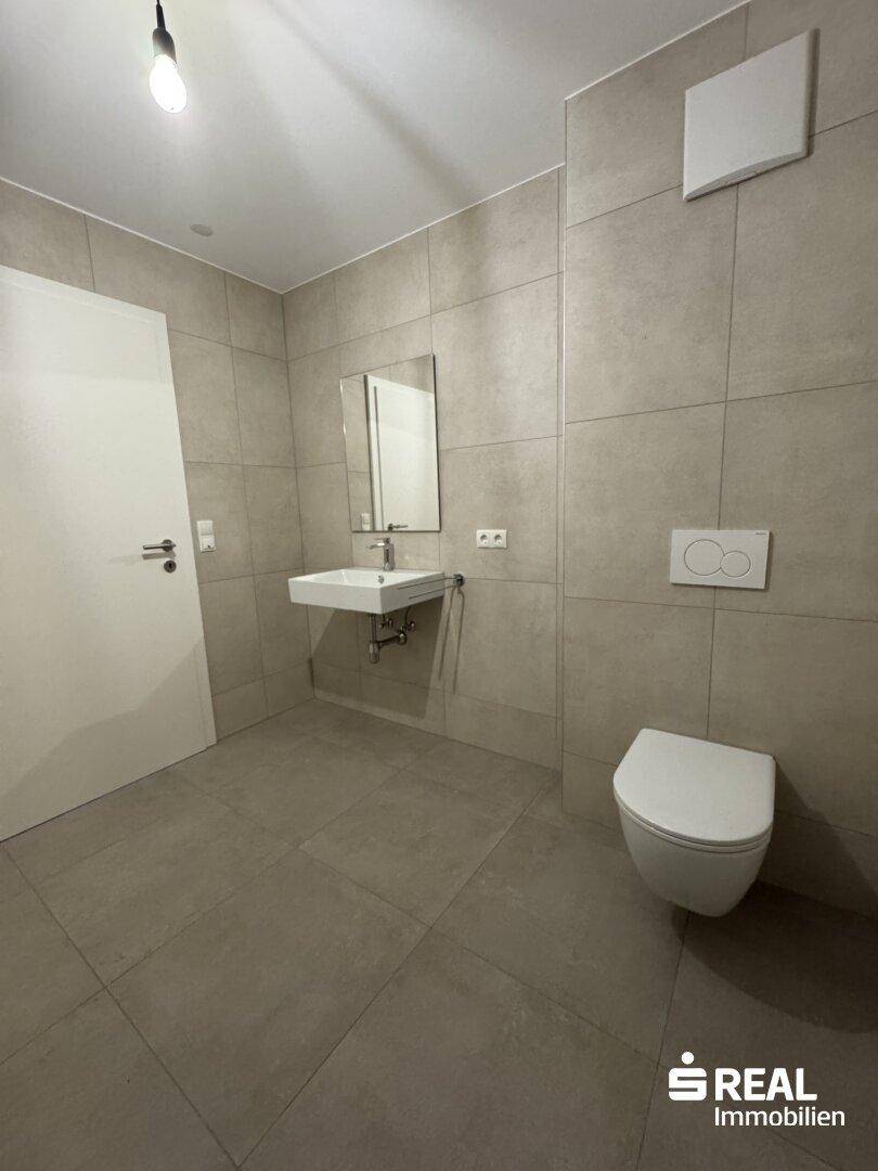 Modernes Badezimmer mit grauen Fliesen, wandhängendem WC und stilvollem Waschbecken mit Spiegel.