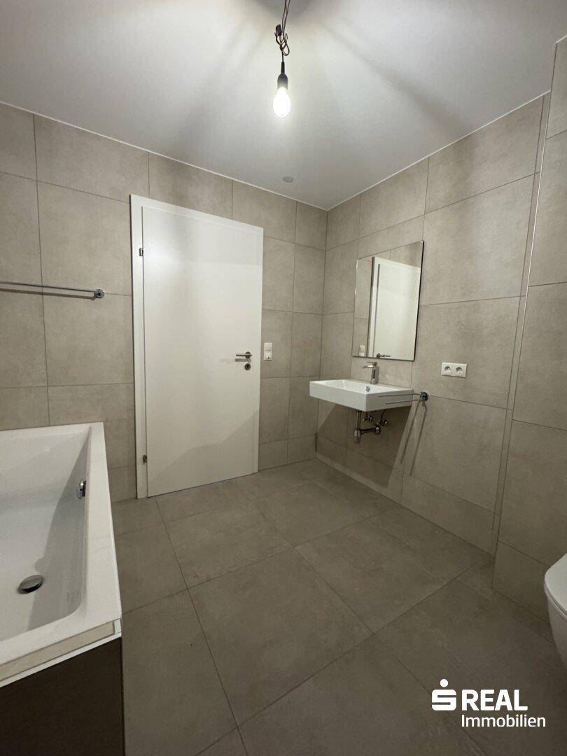 Helles Badezimmer mit modernen grauen Fliesen, Badewanne und wandhängendem Waschbecken.