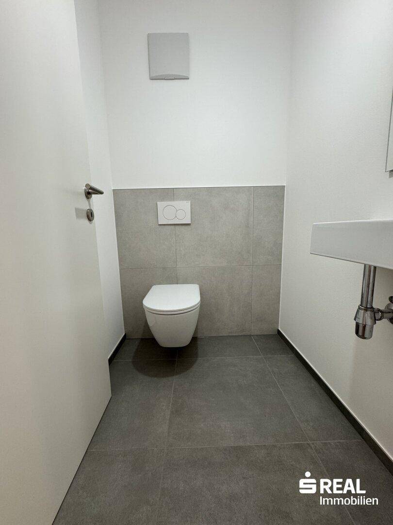 Gäste-WC mit modernen grauen Fliesen, wandhängendem WC und einem einzelnen Waschbecken.