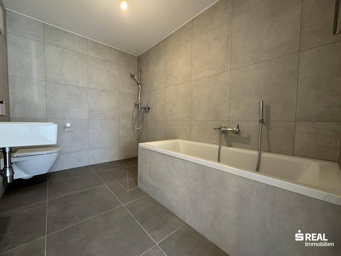 Modernes Badezimmer mit großen grauen Fliesen, einer Badewanne und wandhängendem WC.