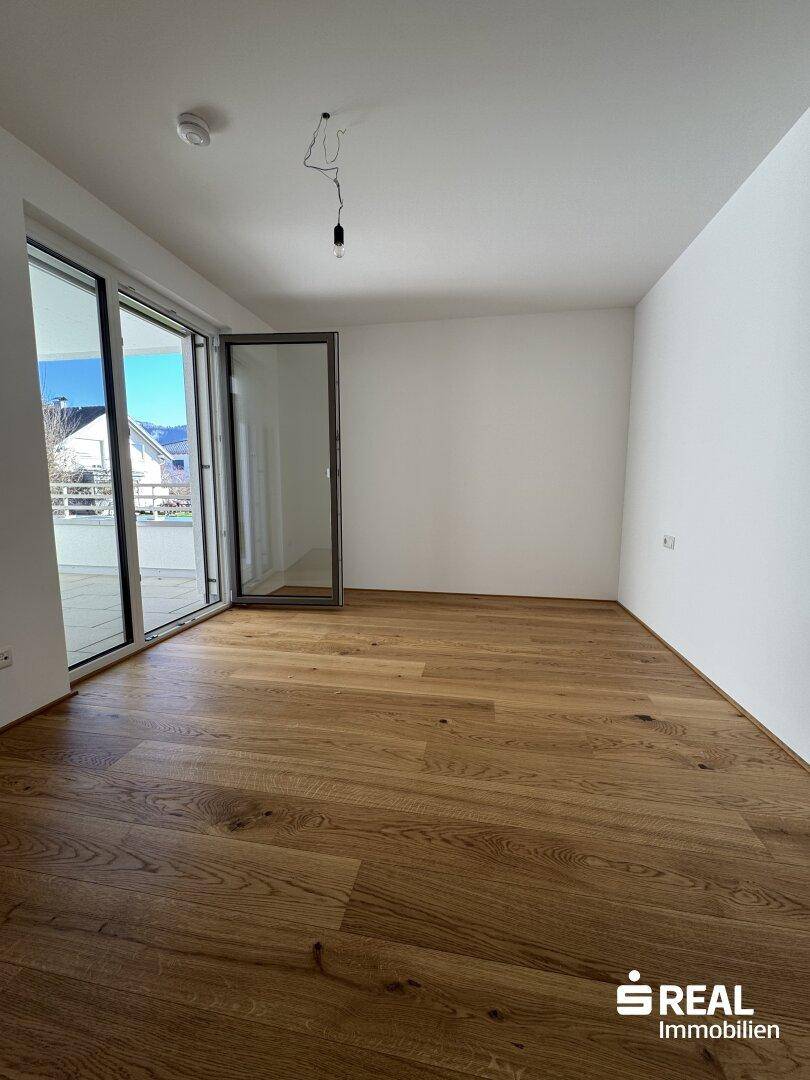 Geräumiges Zimmer mit Holzparkett und großen Glastüren, die Zugang zu einem sonnigen Balkon bieten.