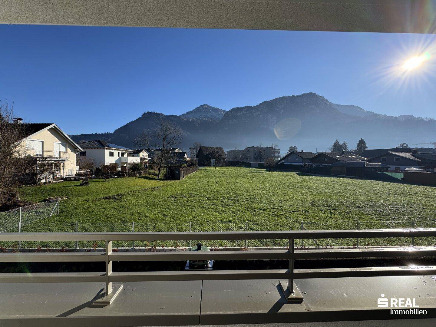 Heller Balkon mit Blick auf grüne Wiesen, umliegende Häuser und die beeindruckende Bergkulisse.
