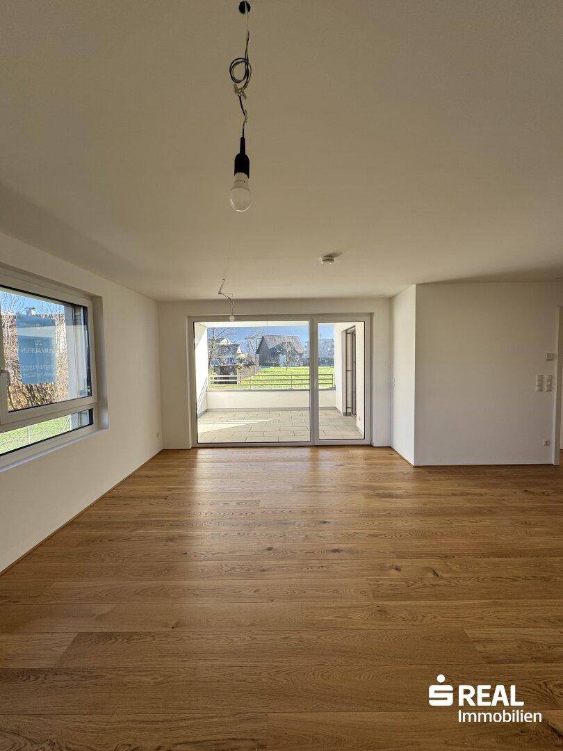 Lichtdurchflutetes Zimmer mit Holzparkett und großen Fenstern, die den Blick nach draußen öffnen.