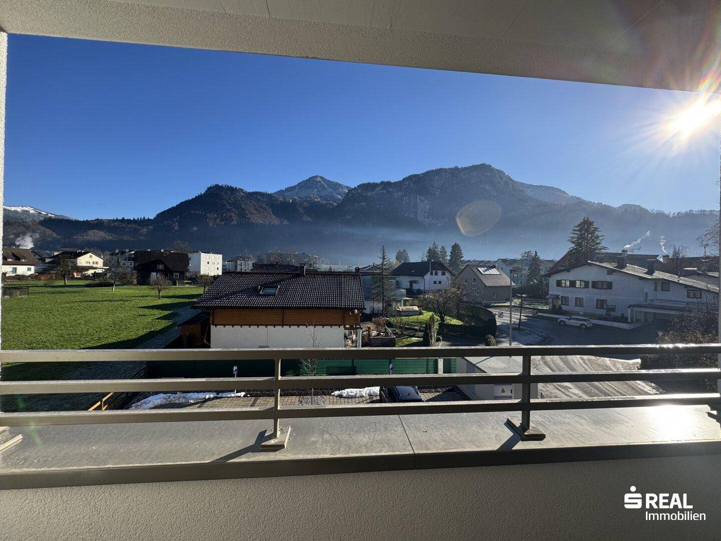 Blick vom Balkon auf die majestätische Bergkulisse und die sonnige Umgebung mit Wohngebäuden.