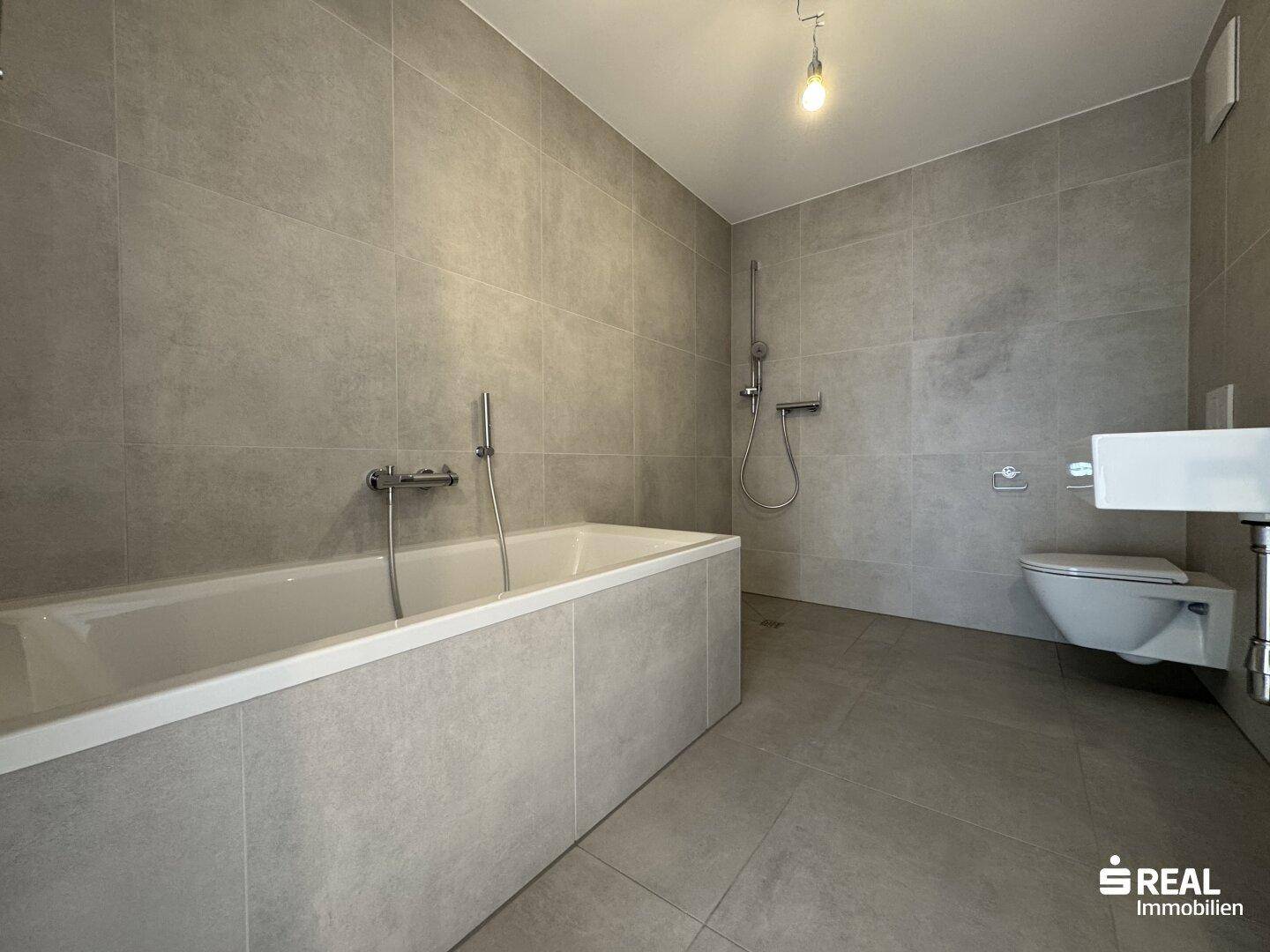 Modernes Badezimmer mit grauen Fliesen, Badewanne mit Duschfunktion und wandhängendem WC.