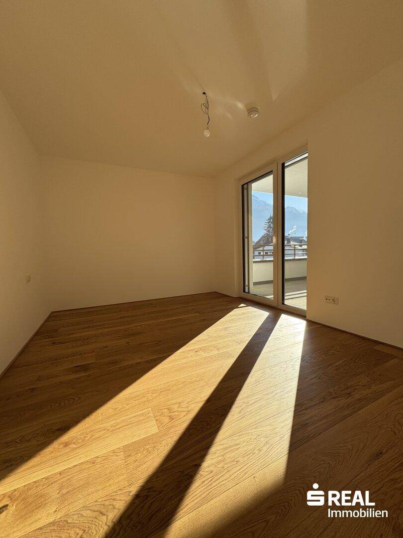 Heller Wohnraum mit Holzboden, großen Fenstern und Zugang zu einem Balkon mit Bergblick.